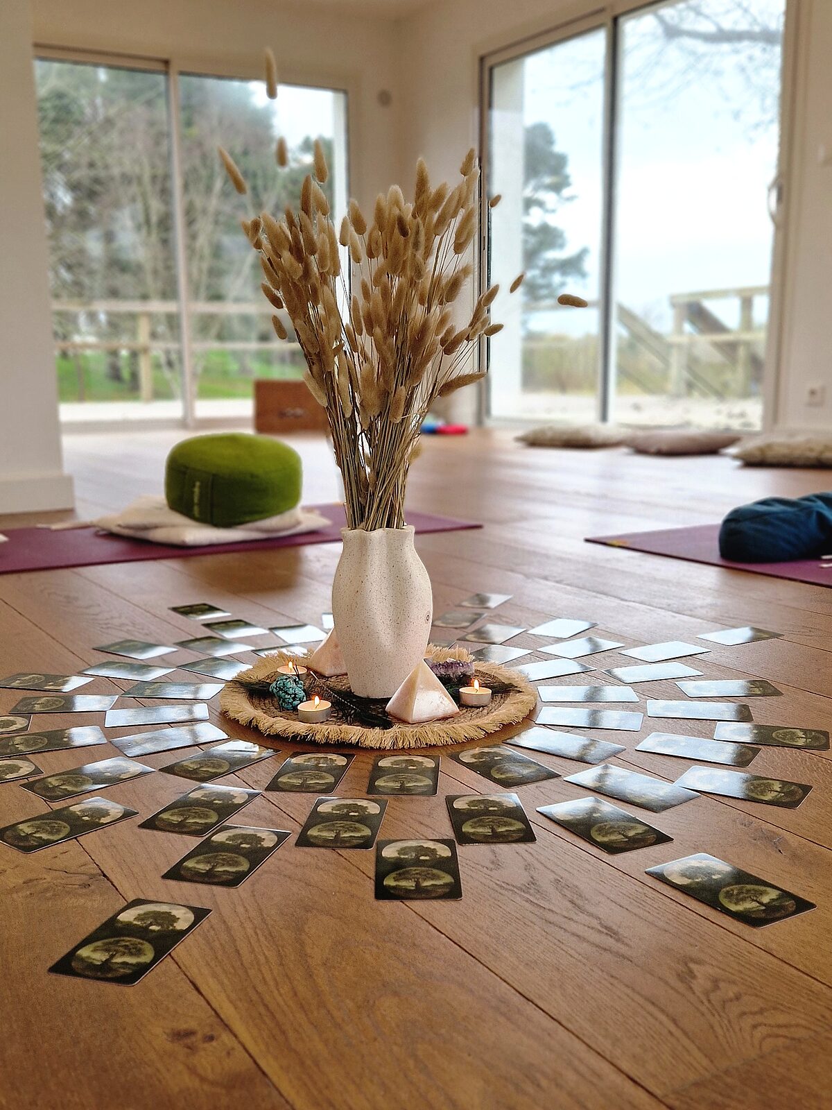 Un large cercle de cartes disposé sur le parquet du cabinet, autour d'un vase de lagurus séchés, de bougies chauffe-plat et de pierres posées sur un tapis de jute ; au fond, des baies vitrées ouvertes sur le jardin, des tapis et des coussins prêts pour une journée de groupe
